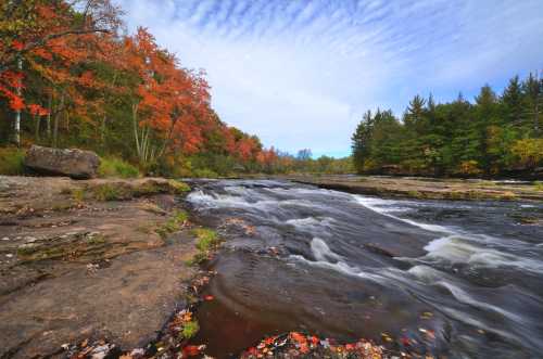 A serene river flows over rocks, surrounded by vibrant autumn foliage and a clear blue sky.