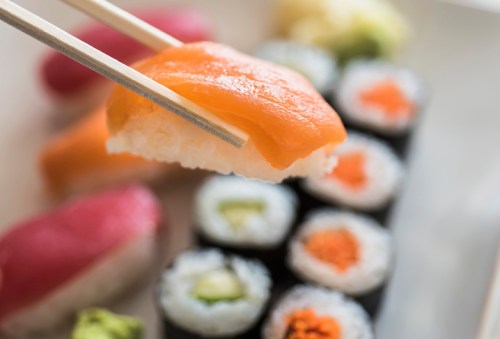 A close-up of sushi being held with chopsticks, featuring salmon on rice and various rolls in the background.