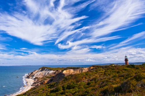 A scenic view of a lighthouse on a cliff, with vibrant blue skies and wispy clouds over the ocean and coastline.