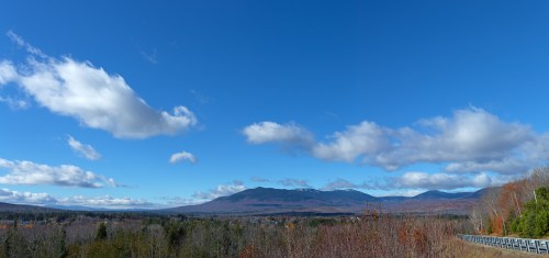 A panoramic view of mountains under a clear blue sky with scattered clouds, surrounded by autumn foliage.