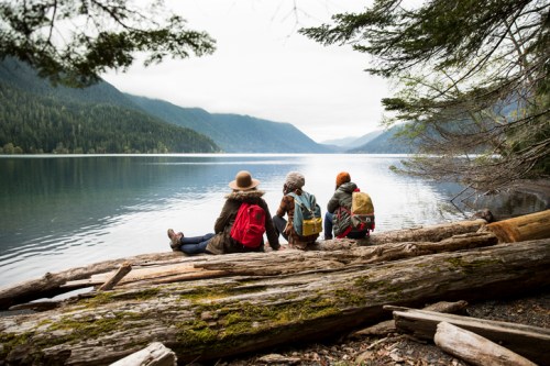 Three hikers sit on a log by a serene lake, surrounded by mountains and trees, enjoying the peaceful view.