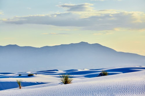 A serene desert landscape with white sand dunes and distant mountains under a soft, cloudy sky.