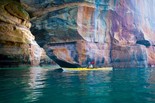 A kayaker paddles near a colorful rock formation, reflecting in calm waters.