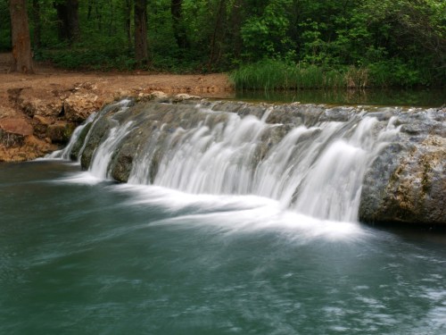 A serene waterfall cascades over rocks into a tranquil, turquoise pool surrounded by lush green trees.