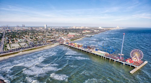 Aerial view of a vibrant pier with amusement rides, waves crashing, and a coastal city in the background.