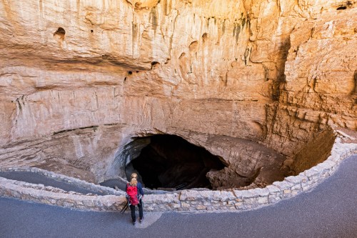 A person stands at the edge of a large cave, gazing into the dark opening surrounded by rocky walls.