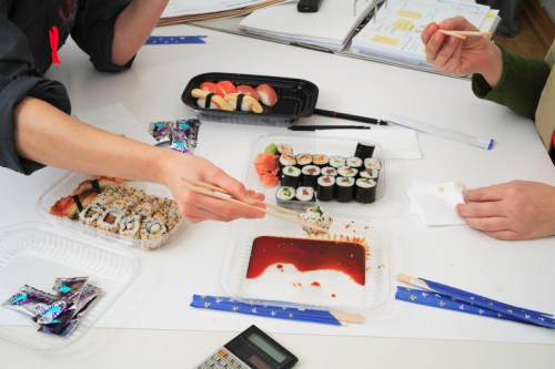Two people sharing sushi at a table, with various sushi rolls and soy sauce in containers. A calculator is visible.