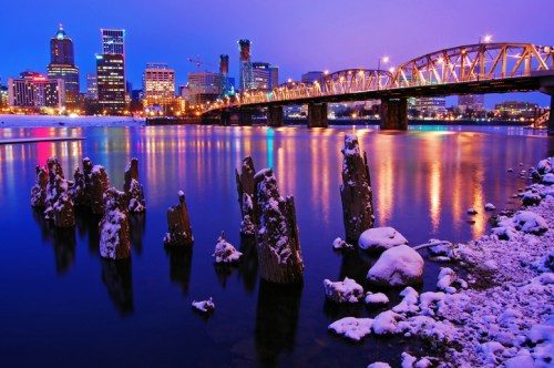 A snowy riverside view of Portland at dusk, featuring a lit bridge and city skyline reflecting in the water.