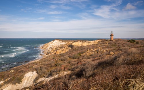 A scenic coastal view featuring a lighthouse on a cliff, with waves crashing against the shore and a blue sky above.