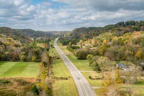 A scenic view of a winding road through lush green fields and rolling hills, with autumn foliage in the background.