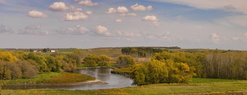 A serene landscape featuring a winding river surrounded by lush trees and rolling hills under a blue sky with clouds.