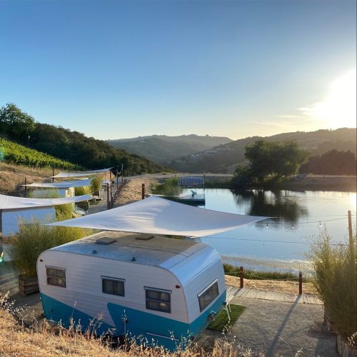 A vintage camper by a serene lake, surrounded by hills and vineyards, with sunlit skies and shade sails overhead.