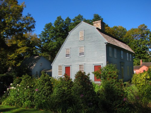 A blue house with red doors surrounded by greenery and trees under a clear blue sky.