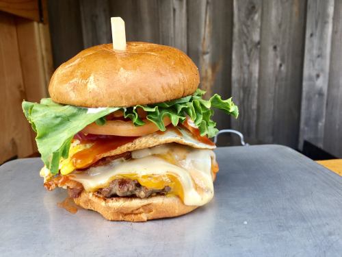 A stacked burger with lettuce, tomato, bacon, cheese, and a fried egg on a toasted bun, served on a metal surface.