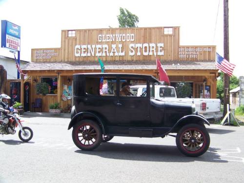 A vintage black car drives past the Glenwood General Store, adorned with flags and rustic wooden architecture.