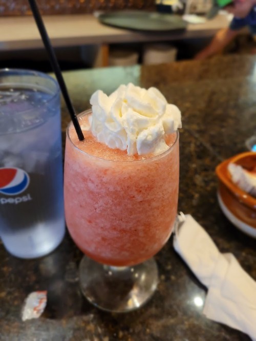 A frosty pink drink topped with whipped cream, served in a glass with a straw, next to a glass of water.