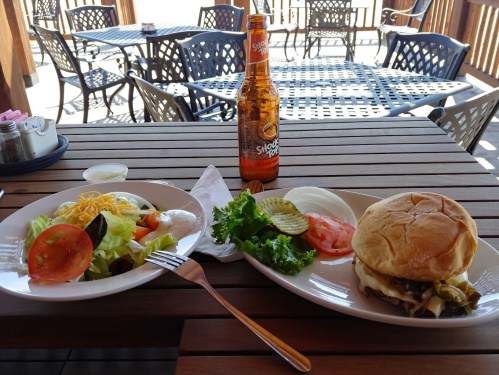 A table setting with a burger, salad, and a bottle of soda, outdoors with chairs in the background.