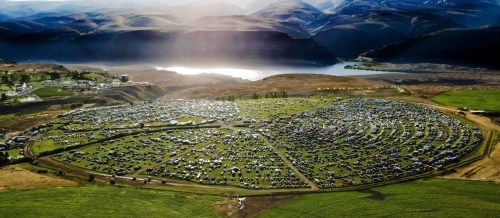 Aerial view of a large circular parking area filled with cars, surrounded by hills and a river in the background.
