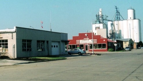 A small town street featuring a restaurant, a service building, and grain silos in the background under a clear sky.