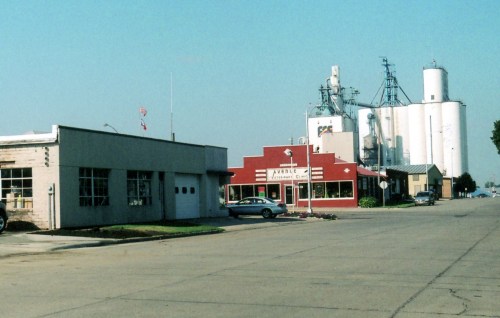 A small town street featuring a red building, a garage, and large silos in the background under a clear blue sky.