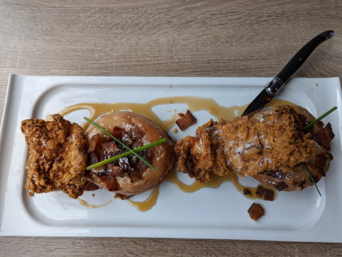 A plate with fried chicken, a glazed donut topped with bacon, and drizzled with syrup, garnished with chives.