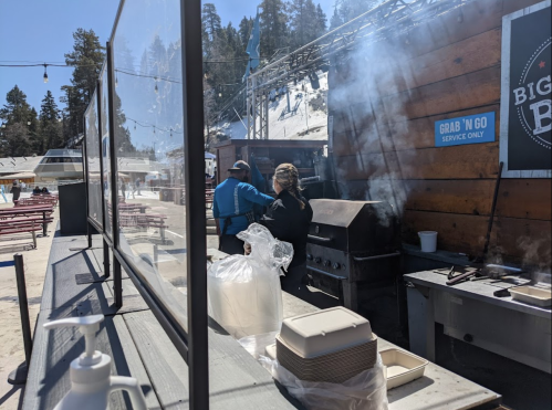 Two people cooking at an outdoor food stand with smoke rising, surrounded by snow-covered trees and seating.