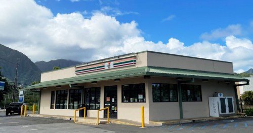 A 7-Eleven store with a green roof, large windows, and a mountain backdrop under a partly cloudy sky.