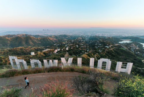A person walks near the Hollywood sign, overlooking a scenic view of Los Angeles at sunset.