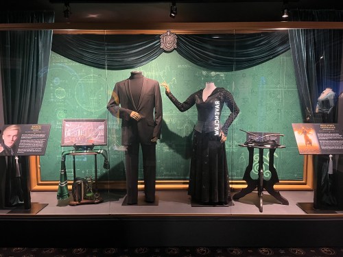 Display case featuring a suit and a black dress, with Hogwarts house items and information panels in the background.