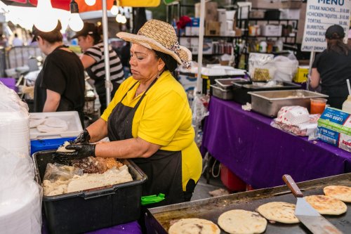 A woman in a yellow shirt and straw hat prepares food at a vibrant market stall, with tortillas cooking on a grill.