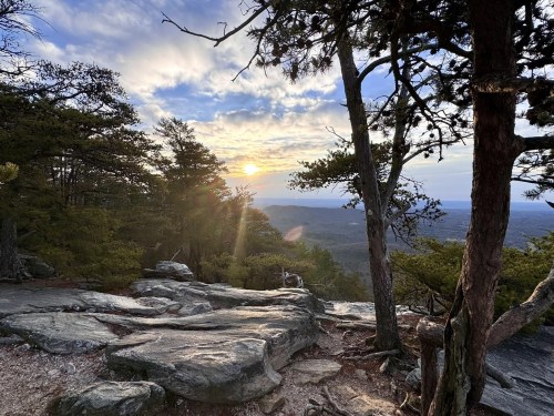 Sunset view from a rocky mountain overlook, framed by trees and clouds in a serene landscape.