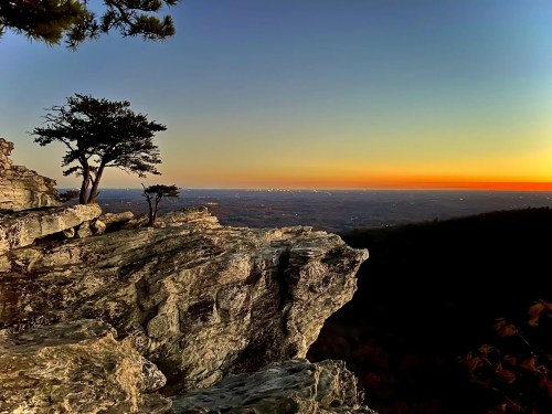 A rocky cliff at sunset, with silhouetted trees and a distant view of a valley illuminated by city lights.