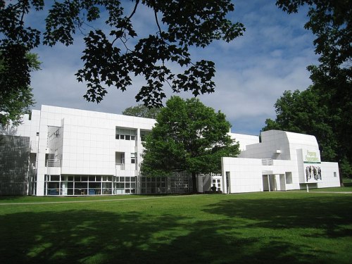 Modern white building surrounded by green grass and trees under a partly cloudy sky.