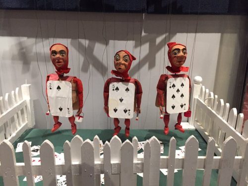 Three marionette puppets dressed in red with playing card motifs, standing behind a white picket fence.