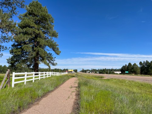 A dirt path lined with grass and a white fence, leading towards a clear blue sky and distant buildings.