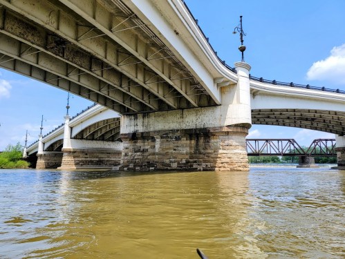 A view from below a large bridge, showcasing its arches and stone pillars over a calm river under a clear blue sky.