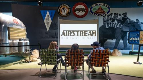 Three people sit in chairs facing a screen displaying "Airstream Heritage Center" in a museum setting.