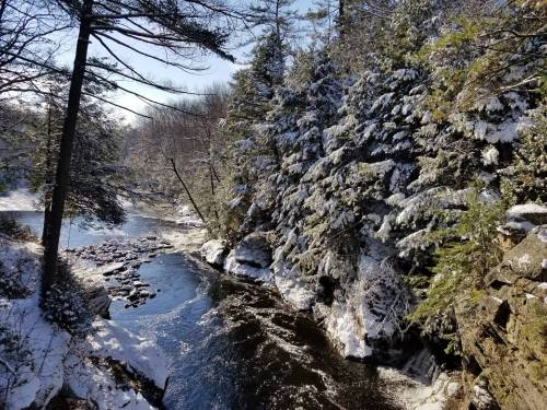 A serene winter scene featuring a river surrounded by snow-covered trees and rocky banks under a clear blue sky.