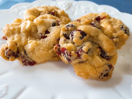 Three freshly baked cookies with cranberries and white chocolate chips on a decorative white plate.