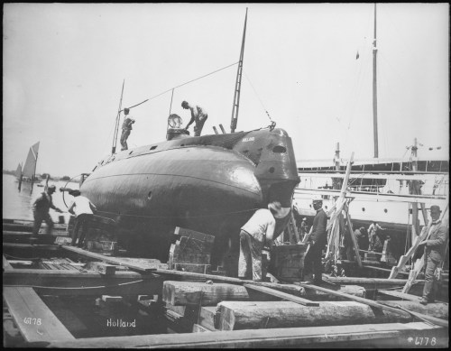 Historic black-and-white photo of workers handling a submarine on a dock, with boats in the background.