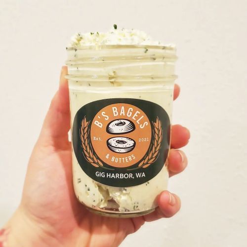 A hand holding a jar of B's Bagels & Butters, featuring a creamy spread with herbs, labeled with the brand and location.