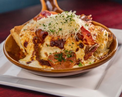 A bowl of cheesy baked pasta topped with crispy bacon and grated cheese, garnished with parsley.