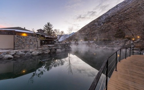 A serene hot spring surrounded by rocks and trees, with mist rising and mountains in the background at dusk.