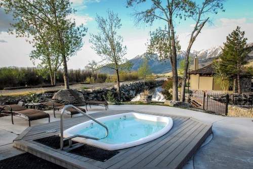 A hot tub on a patio with a scenic view of mountains and trees, beside a flowing stream.