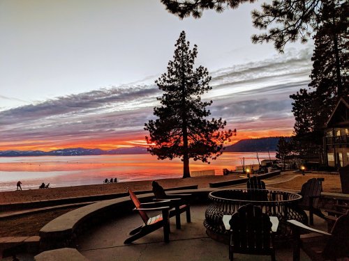 Sunset over a lake with mountains in the background, featuring a fire pit and chairs on a sandy beach.