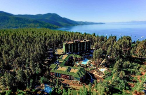 Aerial view of a lakeside resort surrounded by trees and mountains, featuring pools and outdoor amenities.