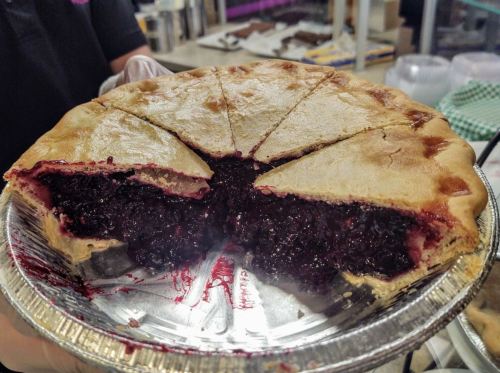 A freshly baked pie with a golden crust and a rich, dark berry filling, partially sliced and displayed in a silver pie tin.