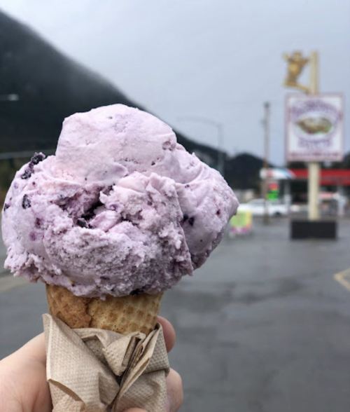 A hand holding a large scoop of purple ice cream in a cone, with a misty mountain and shop sign in the background.