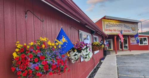 Colorful flower baskets hang outside a red building with signs for "The Huckleberry Patch" gift shop and café.