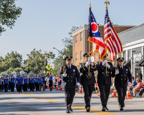 Three uniformed officers march in a parade, carrying the Ohio and American flags, with a marching band in the background.